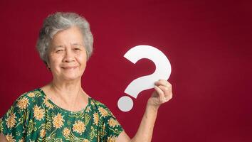 Question mark concept. Senior woman with short gray hair holding a white question mark paper and looking at the camera with a smile while standing on a red background. Space for text photo