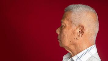 Side view of an elderly Asian man looking away while standing on a red background in the studio. Space for text. Aged people and healthcare concept photo