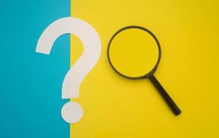 A magnifying glass and a question mark symbol are on blue and yellow background. Search and question mark concept. Top view. Space for text photo