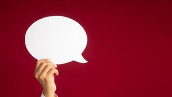 A speech bubble concept. Hand holding an empty white speech bubble over on a red background with copy space for text. Close-up photo