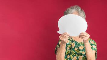 Concept of a speech bubble. A senior woman with short gray hair holding a blank white paper speech bubble closed her face while standing on a red background in the studio. Space for text photo
