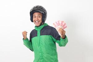 Portrait of Asian online taxi driver wearing green standing against white background, holding a bunch of rupiah money while raising fist, celebrating success photo