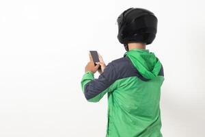 Back view of Asian online taxi driver wearing green jacket and helmet holding mobile phone with blank screen. Isolated image on white background photo