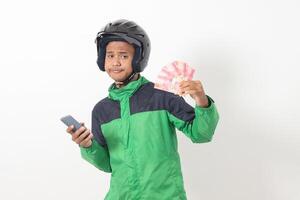 Portrait of Asian online taxi driver wearing green standing against white background, smiling and looking at camera, holding a bunch of rupiah money photo