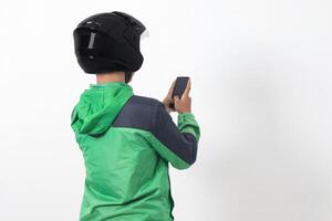 Back view of Asian online taxi driver wearing green jacket and helmet holding mobile phone with blank screen. Isolated image on white background photo