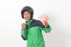 Portrait of Asian online taxi driver wearing green standing against white background, holding a bunch of rupiah money while raising fist, celebrating success photo
