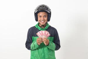 Portrait of Asian online taxi driver wearing green standing against white background, smiling and looking at camera, holding a bunch of rupiah money photo
