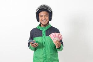 Portrait of Asian online taxi driver wearing green standing against white background, smiling and looking at camera, holding a bunch of rupiah money photo