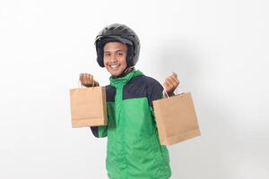 Portrait of Asian online courier driver wearing green jacket and helmet giving craft paper shopping bag with gift inside. Isolated image on gray background photo