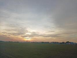 Sunset over a grassy field with distant buildings and trees photo