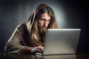 A young handsome monk with long hair sits at a table with a laptop and types on the keyboard. photo