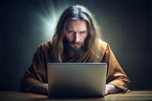 A young handsome monk with long hair sits at a table with a laptop and types on the keyboard. photo