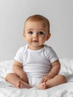 A baby in a white bodysuit sitting on a bed photo