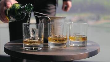A man's hand pours alcohol into a glass with ice. Three glasses of alcohol with ice are not half full, whiskey is added to one of them video
