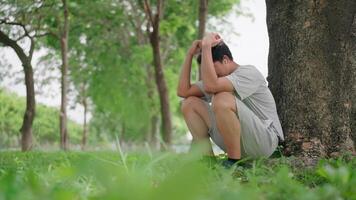 Asian man sits on the ground outside, crying and leaning against a tree, thinking. He thinks about his life, looks forward with hope, and wears AirPods while relaxing in a park. Healthy Lifestyle. video