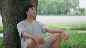 Side view of Asian man leaning on a tree thinking outside sitting on floor, He thinking about life, looking forward with hope while relaxing in a park, Healthy lifestyle. video