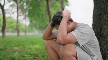 Side view of Asian man sitting on the ground outside crying and leaning against a tree. He is contemplating life, looking ahead with hope, and wearing Air Pods while relaxing in a park. video