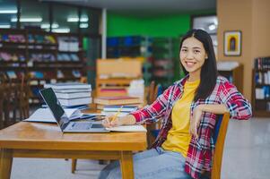 Smiling female student posing while working on laptop and searching for books to study, make report, find useful information in university room. photo