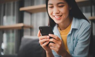 Smiling young asian woman using mobile phone checking social media,app playing game, shopping online, ordering delivery relax while sitting on a couch at home with laptop computer photo