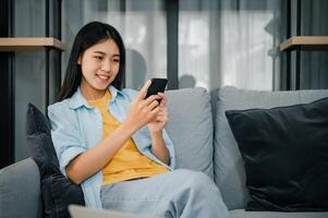 Smiling young asian woman using mobile phone checking social media,app playing game, shopping online, ordering delivery relax while sitting on a couch at home with laptop computer photo