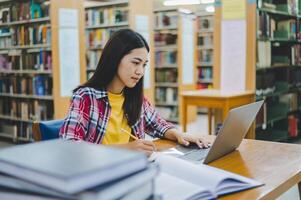 Female college student working on laptop and searching for books to study, make report, find useful information in college room. Concept of reading, learning and intelligence. photo