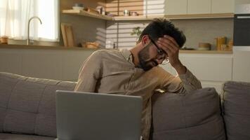 Shocked upset Indian Arabian man guy business work from home kitchen negative bad result worried anxious male freelancer on couch sofa read laptop notification computer problem fail difficult crisis photo