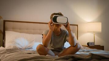 Little modern Caucasian child wearing vr glasses virtual reality headset boy son bed playing online 3d virtual game kid exploring digital universe world game alone evening illuminated bedroom indoors photo