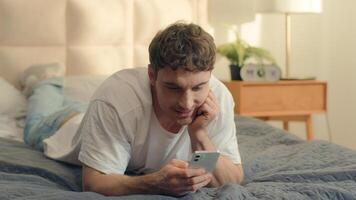 Caucasian man lying on bed using mobile phone smartphone cellphone texting smiling guy male communication online web technology internet social media networks bedroom home enjoy sms chatting gadget photo