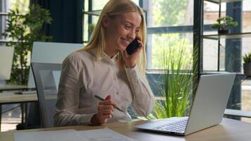 Happy Caucasian businesswoman talk phone conversation consult client business commerce in office smiling woman girl multitask working use laptop check documents on desk support customer mobile call photo