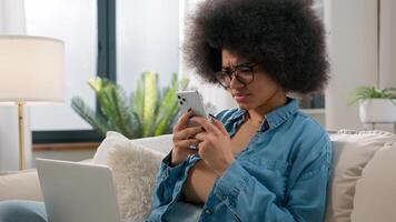African American woman ethnic girl freelancer with laptop on couch browsing mobile phone take break in distant work remote e-learning computer using scrolling smartphone app read confused news at home photo