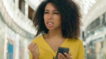 Young unhappy African American woman looking at screen display smartphone telephone error failure evil angry female screaming problem trouble breaking dissatisfied bad news messages annoyed technology photo