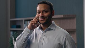 American male office worker at workplace in corporate company businessman talking mobile phone smiling multiracial man speak business negotiate discuss answer call smiling at camera talk smartphone photo