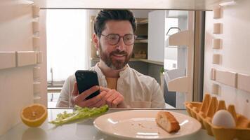 Man hungry guy open empty refrigerator with half lemon, nasty lettuce leaf, one egg, slice of bread order food delivery buying products with mobile phone deliver app point of view from inside fridge photo