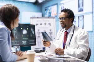 Professional medic analyzing x ray imaging and test findings in office, giving the diagnostic report for female patient at check up. African american specialist examines scans for interpretation. photo