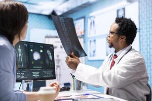 Professional medic analyzing x ray imaging and test findings in office, giving the diagnostic report for female patient at check up. African american specialist examines scans for interpretation. photo