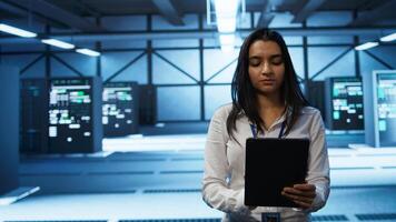 Software developer walking in data center, using tablet to design, implement and manage gear, ensuring optimal performance and reliability. IT worker in server farm coding on device, camera B photo