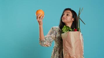 Indian woman with paper bag with vegetables and fruits testing quality, looking at orange, studio background. Vegetarian verifying groceries are ripe after buying them from zero waste shop, camera A photo