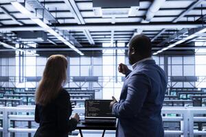 Manager overseeing data center next to employee, doing yearly infrastructure inspection. Coworkers doing brainstorming in server farm, monitoring supercomputers equipment with laptop photo
