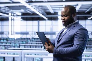 System administrator using tablet in data center with advanced tech, ensuring cybersecurity and efficient network management. Man in server hub optimizing data center equipment with device photo