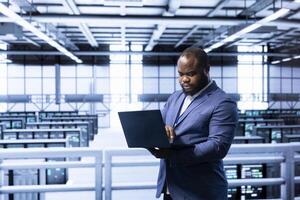 Software developer in data center implementing backup solutions to protect against data loss using laptop, ensuring efficiency. High tech workspace specialist resolving technical issues using notebook photo