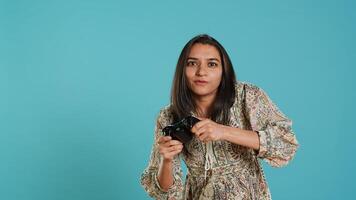 Gamer annoyed after losing online multiplayer videogame, being defeated by rival players. Indian woman upset after seeing game over message, holding controller, studio background, camera A photo
