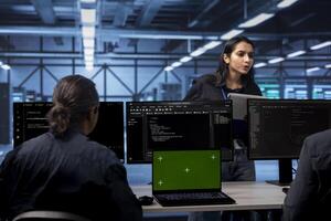Multiethnic team in server hub ensures data remains shielded from potential threats using green screen laptop. Diverse colleagues safeguarding data center against malicious access with mockup notebook photo