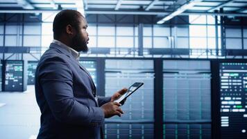 Technician walking in server room, doing checkup, inputting data on tablet of needed fixes. IT professional working in data center facility housing storage infrastructure, camera B photo