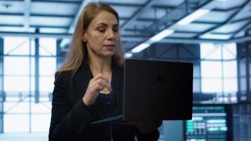 Engineer doing yearly checkup to prolong data center electronics life span and avoid downtime. Woman in server room monitoring server infrastructure components using laptop, camera A photo