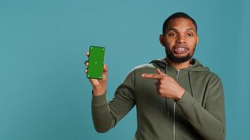 Portrait of african american man doing influencer marketing using green screen phone, studio background. BIPOC person holding empty copy space mockup smartphone device, camera A photo