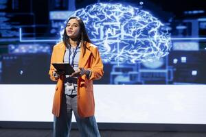 Happy executive holding tablet speaking at event bringing together leading researchers to explore developments in AI and machine learning. Jolly engineer using device to hold tech presentation photo