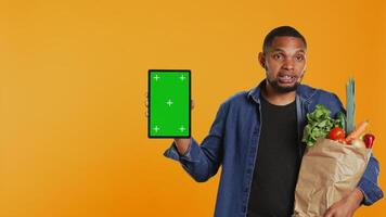 Vegan adult holding a tablet with greenscreen layout in studio, carrying paper bag full of ethically sourced chemicals free produce. Guy advocating for healthy eating with mockup screen. Camera B. photo