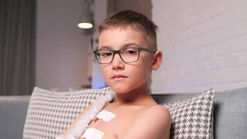 Boy in glasses with sad eyes looks at camera. Holter connected to child at home video