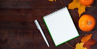 Hello autumn concept. Fallen yellow leaves, pumpkins and a blank notepad on a wooden background. photo