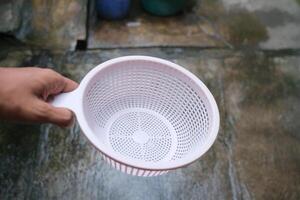 A hand holds a white plastic colander over a gray surface. photo